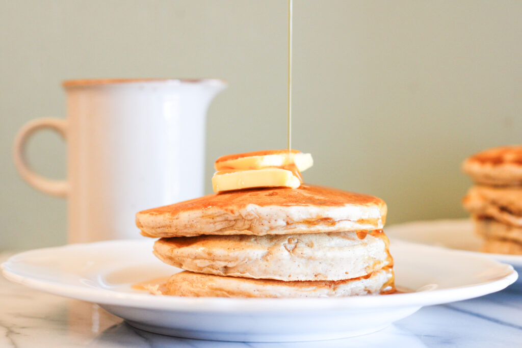 3 banana pancakes on white plate with 2 pats of butter on top and syrup pouring down from above.