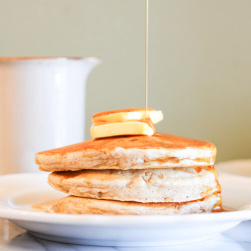 3 banana pancakes on white plate with 2 pats of butter on top and syrup pouring down from above.