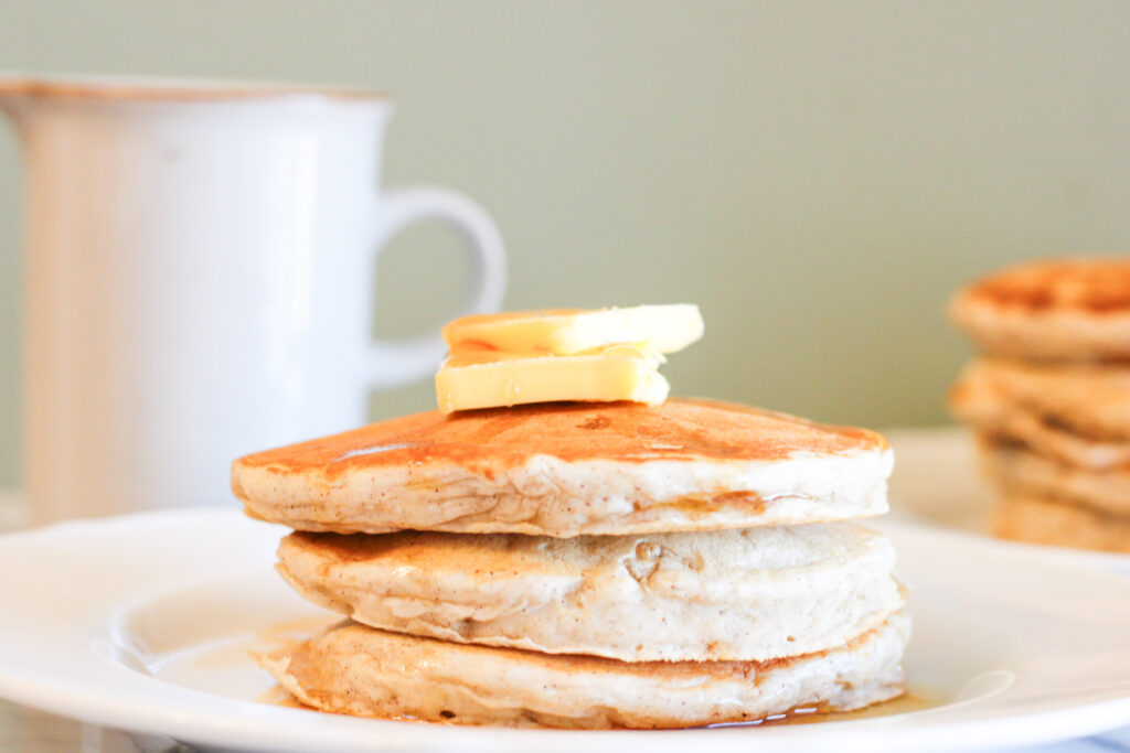 3 banana pancakes on white plate with 2 pats of butter on top and syrup dripping down sides.