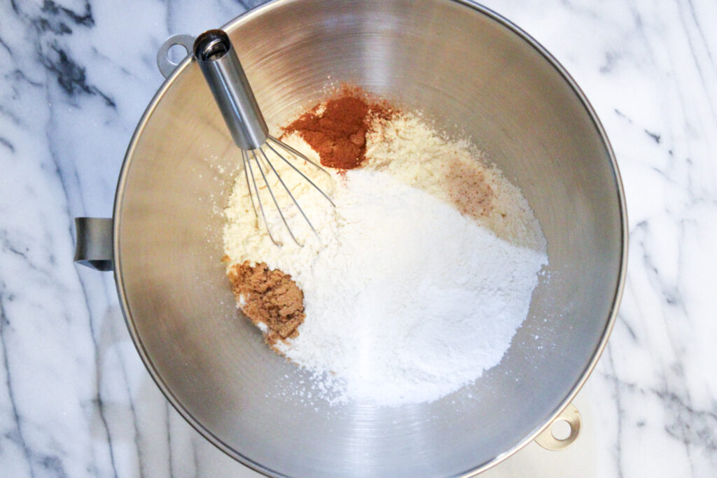 Silver mixing bowl on marble countertop with flour, brown sugar, cinnamon, salt, and wire whisk all in bowl.