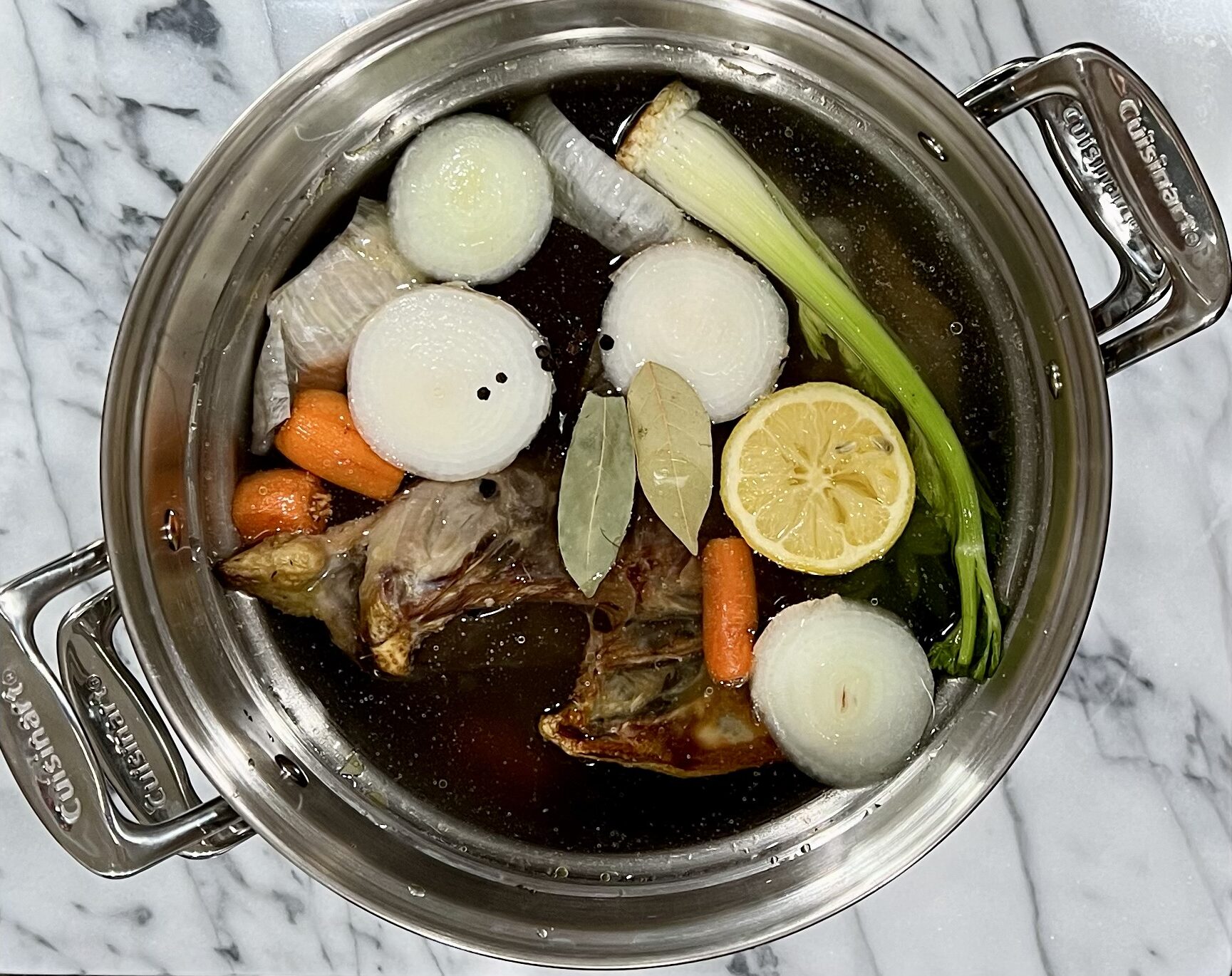 Stainless steel stock pot on marble counter top with celery, onion, lemon, carrots, peppercorns, chicken bones, and 2 bay leaves.