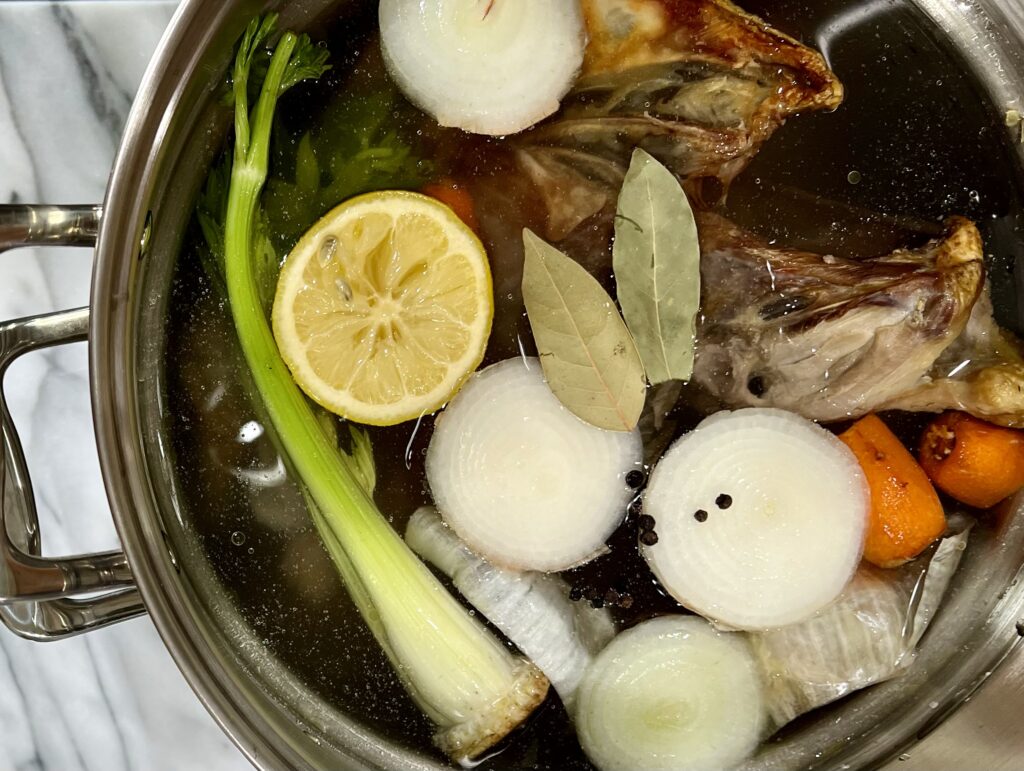 Overhead view of stainless steel stock pot containing carrot tops, lemon, celery, onions, bay leafs, and chicken bones. 