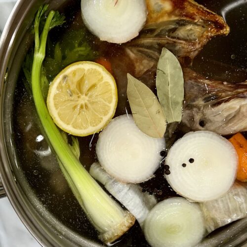 Overhead view of stainless steel stock pot containing carrot tops, lemon, celery, onions, bay leafs, and chicken bones.