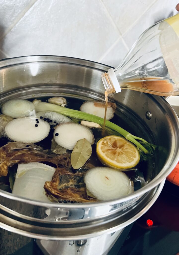 Apple cider viengar being poured into stainless steel stock pot containing carrot tops, lemon, celery, onions, bay leafs, and chicken bones. 