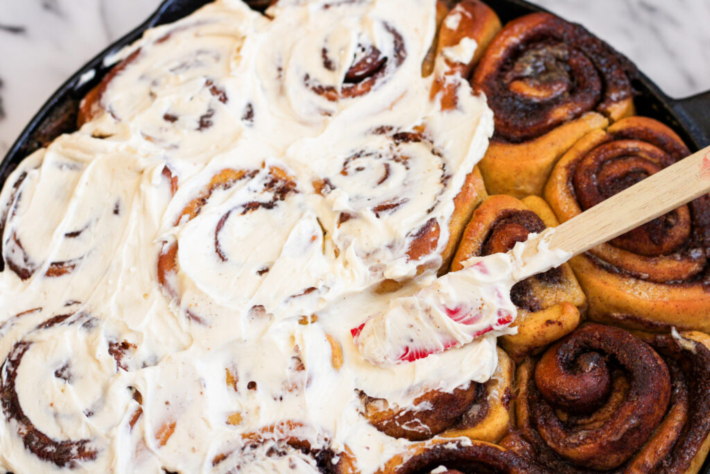 Baked and being frosted Pumpkin Spice Cinnamon Rolls in Cast Iron Pan.