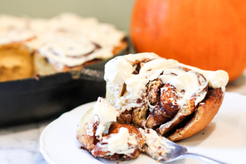 Pumpkin Cinnamon Roll with bite taken out sitting on plate with fork. Pumpkin and cast iron pan with cinnamon rolls in background. 