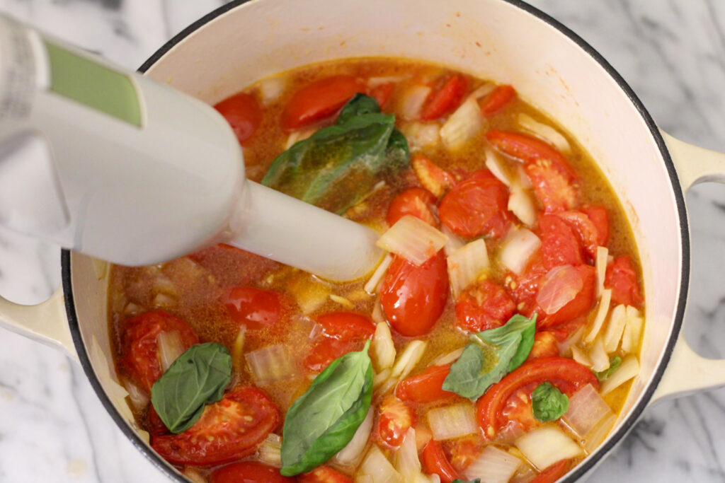 Immersion blender in pot of tomatoes, basil, and onion.