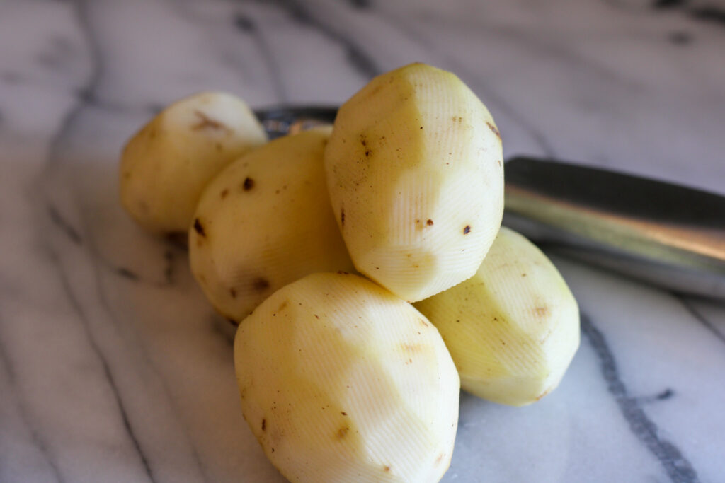 Peeled potatoes on marble countertop with peeler in background. 