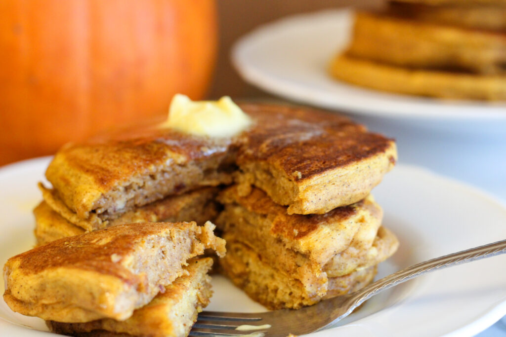 Pumpkin Pancakes with bite missing, butter on top and pumpkin in background. 