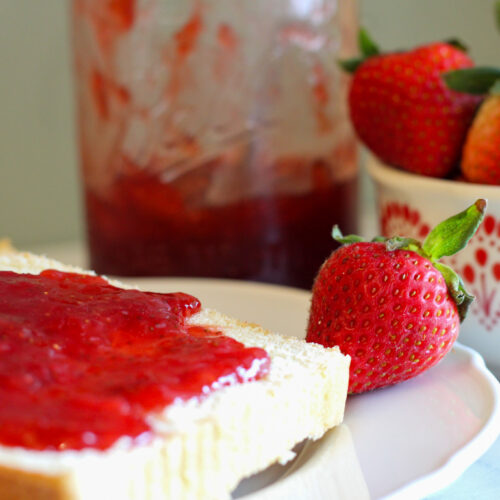 Strawberry jam on slice of bread on white plate. Bowl of strawberries and jar of jam in background.