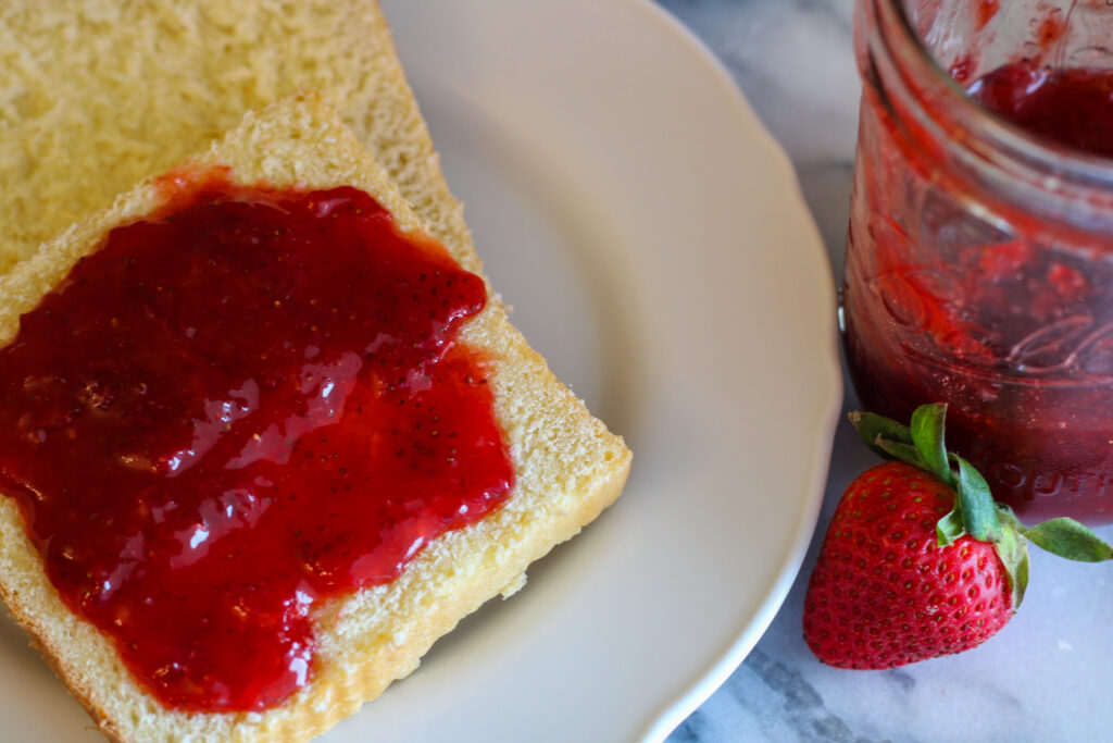 Strawberry jam on slice of bread on white plate. Jar of jam and fresh strawberry to the side.