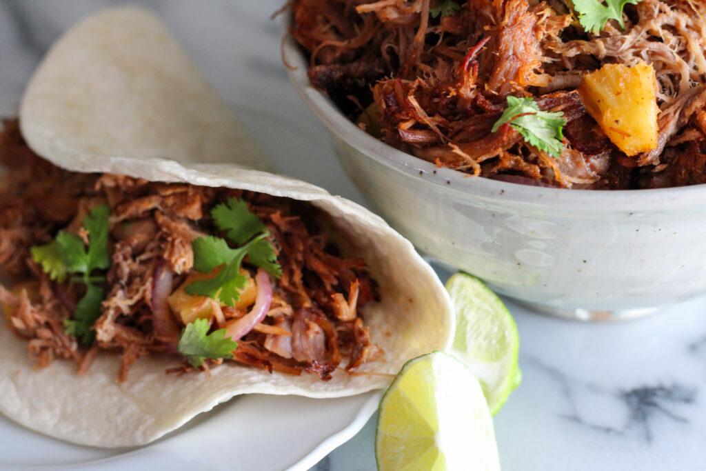 Bowl of tacos al pastor meat with shredded pork taco on plate with wedge of lime.