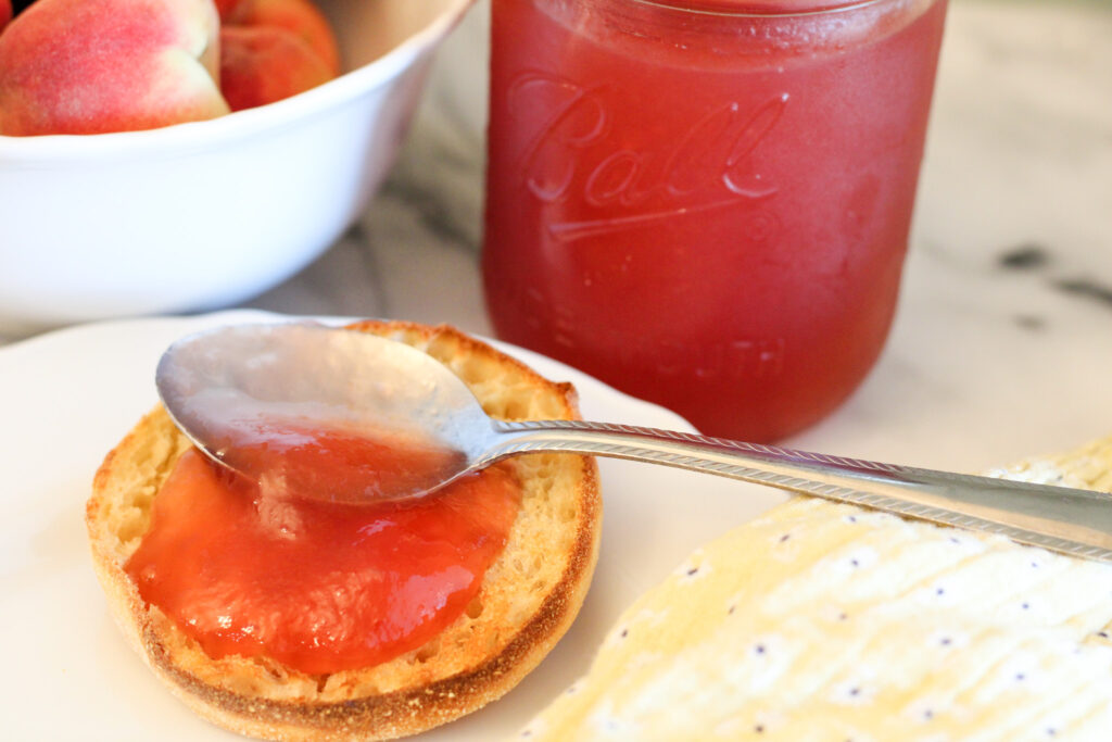 Bowl of peaches, jar of peach jam, and English muffin with peach jam on white plate.
