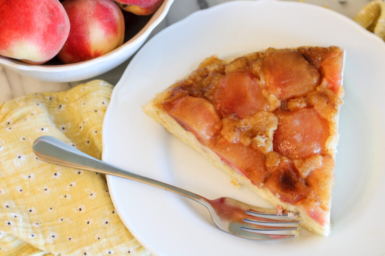 Slice of peach upside down cake with fork and bowl of peaches in back.