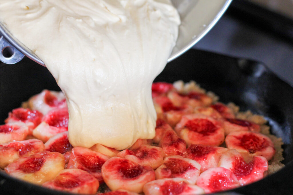 Cake batter being poured over peaches in cast iron pan.