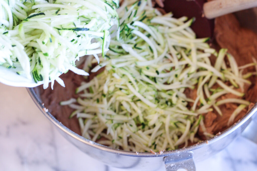 Shredded zucchini being poured into stainless steel bowl of chocolate batter.