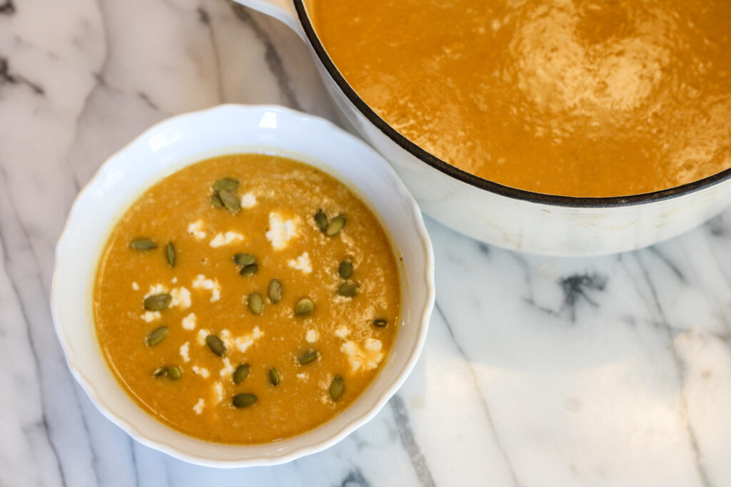Bowl of butternut squash soup with pumpkin seeds and goat cheese next to pot of butternut squash soup.