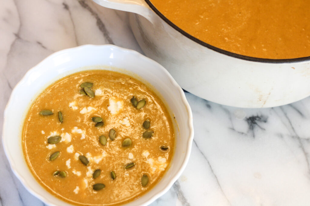 Bowl of butternut squash soup with pumpkin seeds and goat cheese next to pot of butternut squash soup.