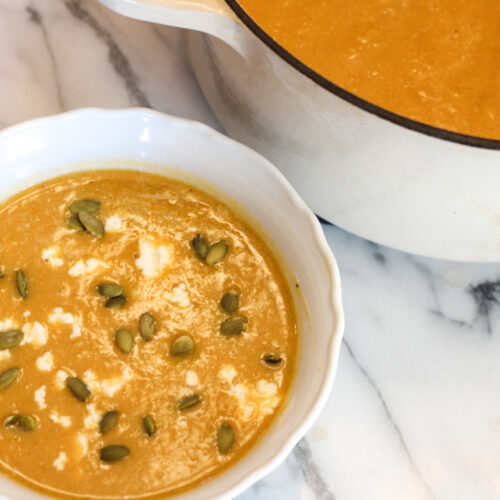 Bowl of butternut squash soup with pumpkin seeds and goat cheese next to pot of butternut squash soup.
