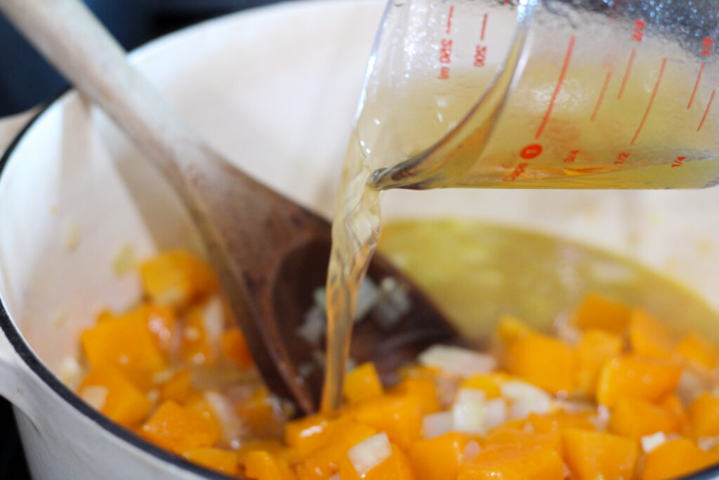 Pouring chicken broth into pot with butternut squash and onions.