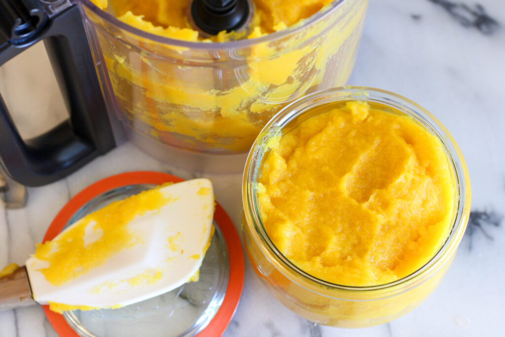 Open jar of pumpkin puree next to food processor and spatula.