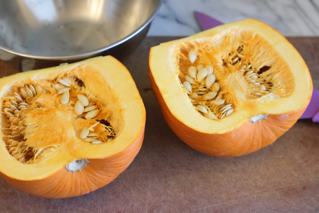 Pumpkin halves face up on cutting board with purple knife and stainless steel bowl.