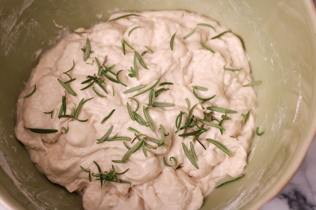 Sourdough bread dough in bowl with fresh rosemary sprinkled on top.